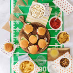 An overhead flat-lay of a football-themed party spread on a green field print. The centerpiece is a wooden board with mini sliders, surrounded by bowls of popcorn, salsa, tortilla chips, nuts, and drinks, with bunting and patterned napkins on a white marble surface.