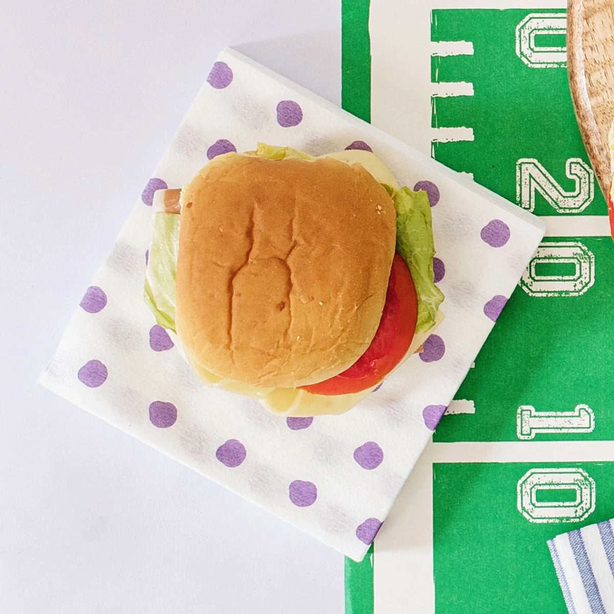 A top-down view of a hamburger on a white paper napkin with purple polka dots, placed on a green football-themed paper mat with white yard-line markings.