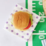A top-down view of a hamburger on a white paper napkin with purple polka dots, placed on a green football-themed paper mat with white yard-line markings.