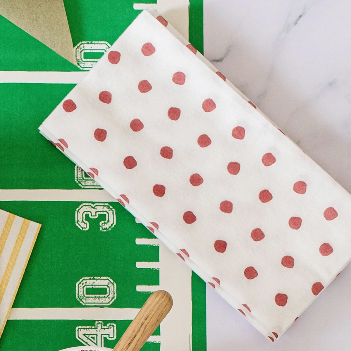 A folded white fabric item, likely a napkin or small towel, printed with a pattern of red polka dots, laid diagonally on a green football field print background.