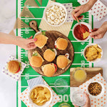 People preparing food on a football-themed tablecloth with burgers, chips, and drinks.
