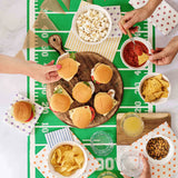 People preparing food on a football-themed tablecloth with burgers, chips, and drinks.