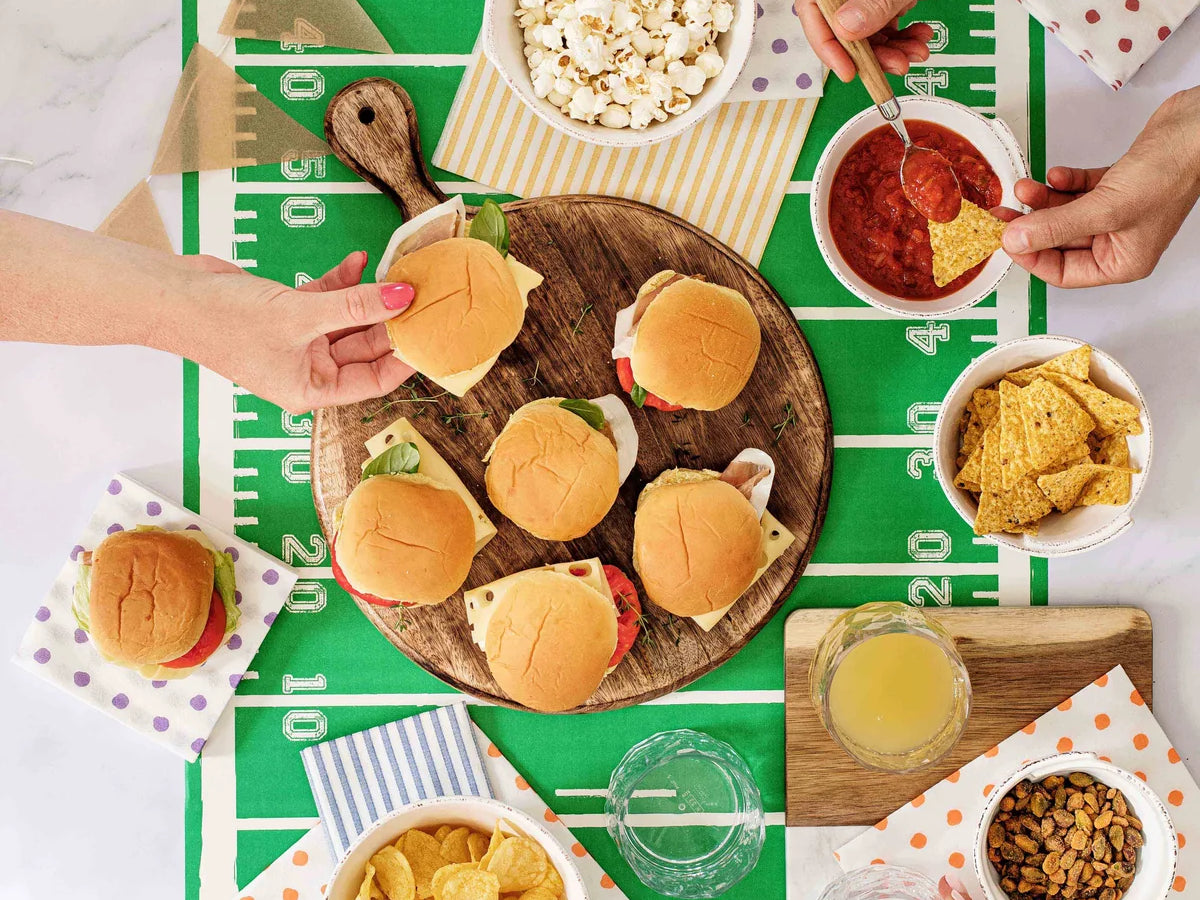 People preparing food on a football-themed tablecloth with burgers, chips, and drinks.