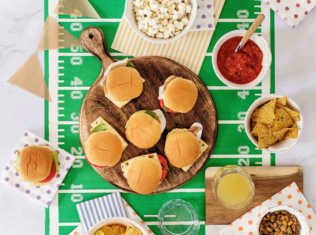 An overhead flat-lay of a football-themed party spread on a green field print. The centerpiece is a wooden board with mini sliders, surrounded by bowls of popcorn, salsa, tortilla chips, nuts, and drinks, with bunting and patterned napkins on a white marble surface.