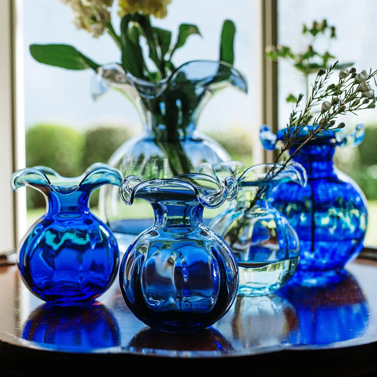 A set of cobalt blue and clear glass flower vases arranged on a dark wooden table in front of a bright window.