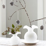 Decorative setup with white vases, greenery, and hanging pinecones on a neutral background