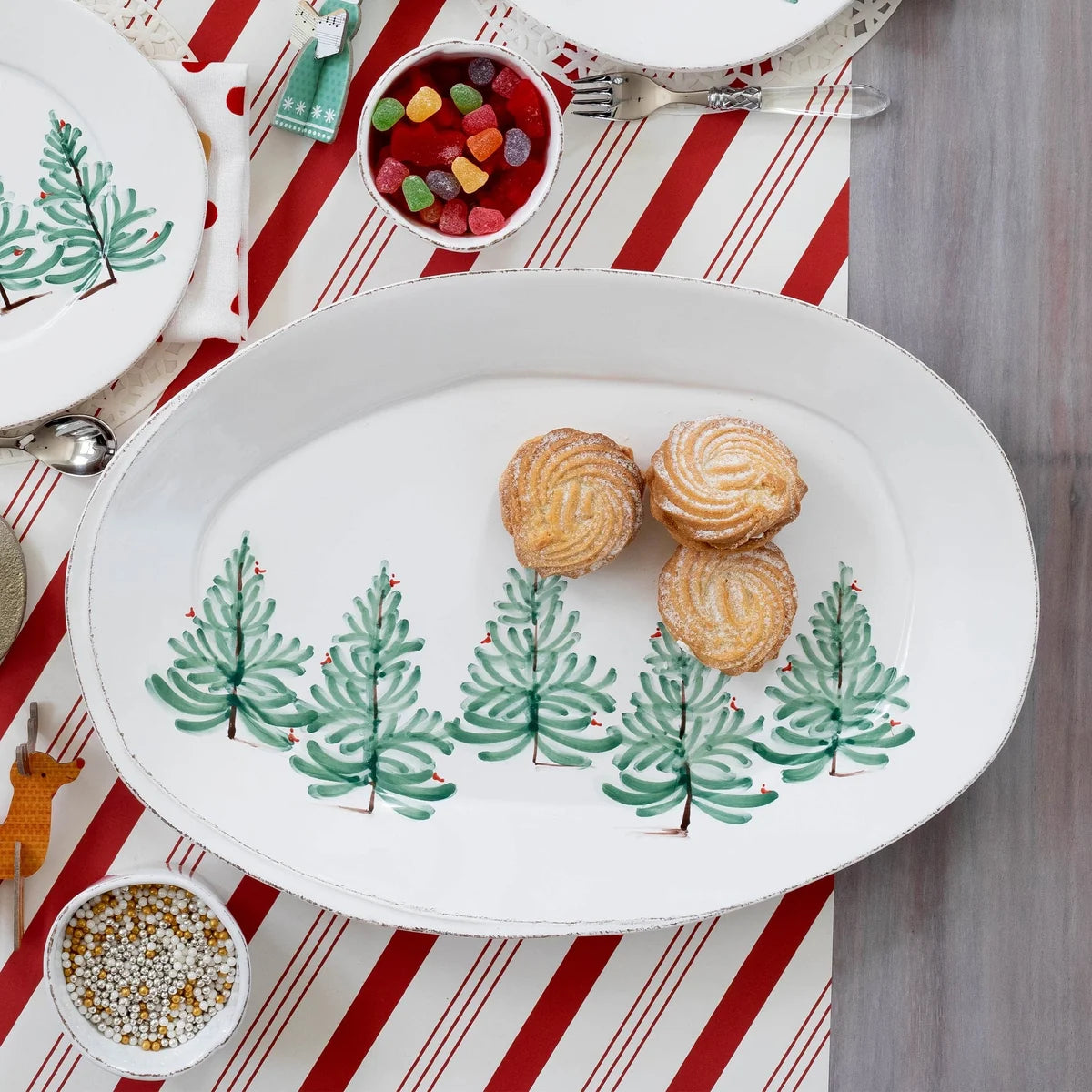 An oval white serving platter decorated with a row of green Christmas trees, styled on a red and white striped table runner with cookies and small bowls of candy and sprinkles.