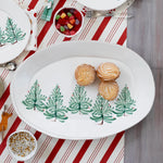 An oval white serving platter decorated with a row of green Christmas trees, styled on a red and white striped table runner with cookies and small bowls of candy and sprinkles.