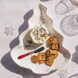 A white ceramic Christmas tree-shaped serving platter styled with cheese, crackers, and a small knife, photographed from above on a light surface with decorative snowflake overlays and shadows.