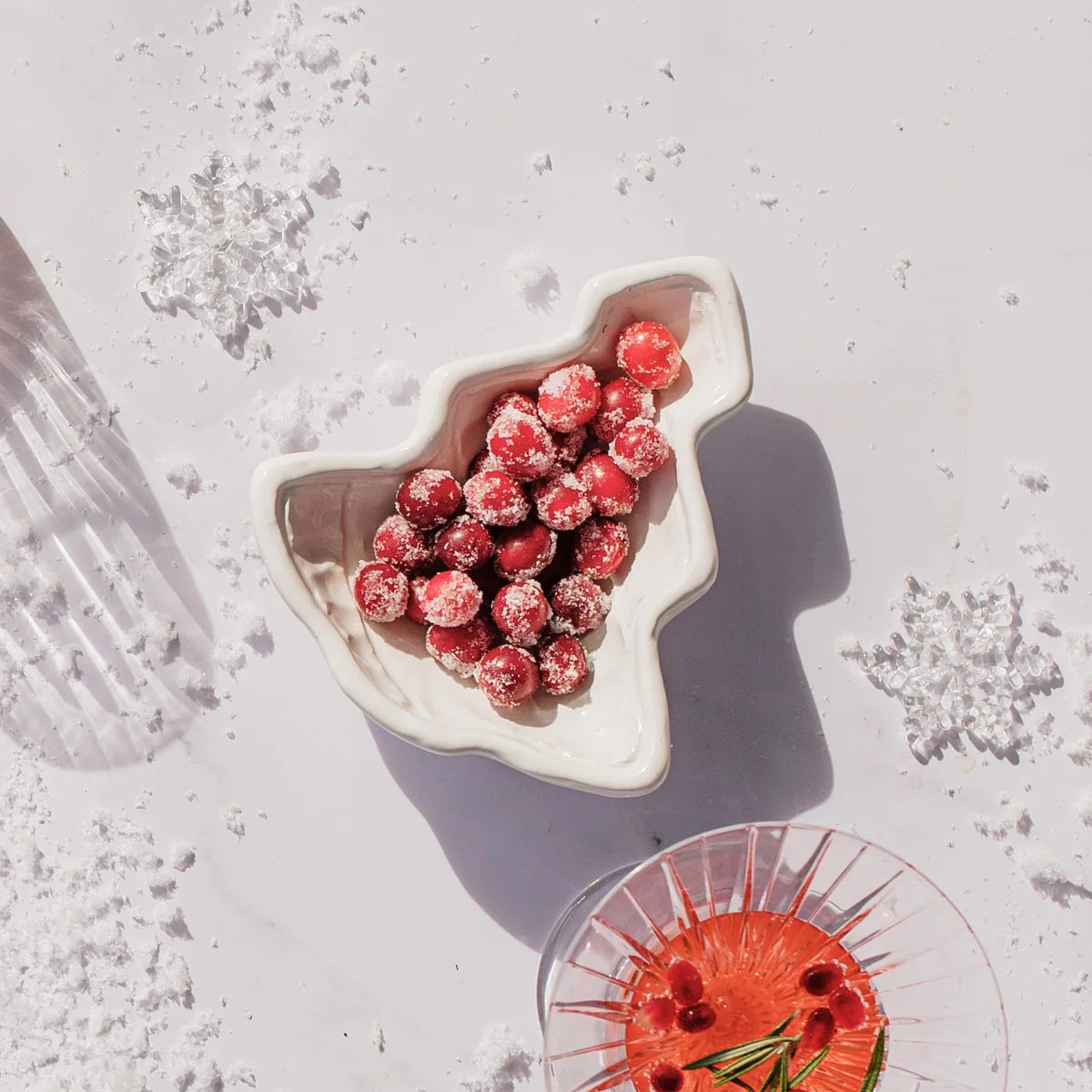 A white ceramic Christmas tree-shaped bowl filled with sugar-dusted red cranberries, styled on a white surface with snowflake decorations and a glass of cranberry juice in the foreground.