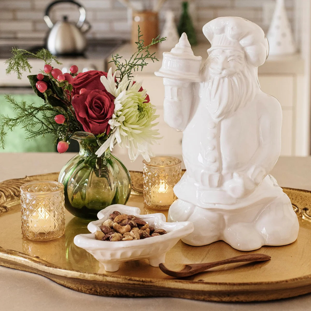 A white ceramic Santa Claus figurine holding a small tower, displayed on a gold tray beside a green glass vase with red and white flowers, lit candle holders, and a small dish of nuts in a kitchen setting.