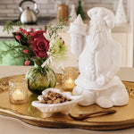 A white ceramic Santa Claus figurine holding a small tower, displayed on a gold tray beside a green glass vase with red and white flowers, lit candle holders, and a small dish of nuts in a kitchen setting.