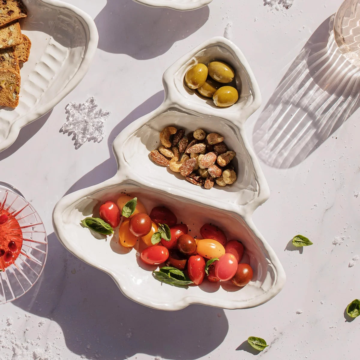 A white ceramic holiday tree-shaped serving tray holding olives, nuts, and cherry tomatoes, styled on a light marble surface with other food props.