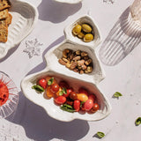 A white ceramic holiday tree-shaped serving tray holding olives, nuts, and cherry tomatoes, styled on a light marble surface with other food props.