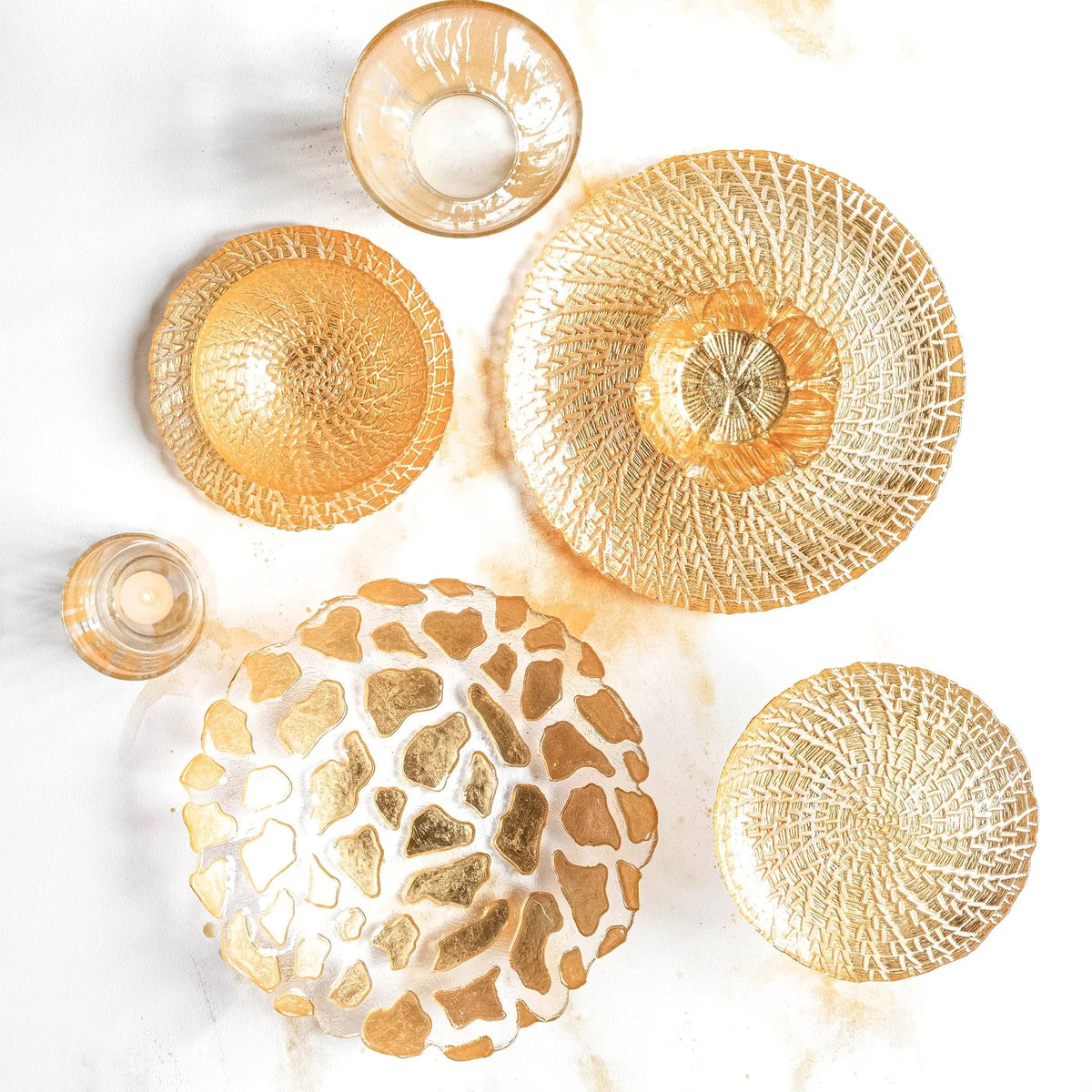 Five gold glass tableware pieces arranged on a white surface: two shallow round plates, two textured bowls, and one small tumbler. The pieces have organic cracked-glass or web-like patterns in metallic gold tones.